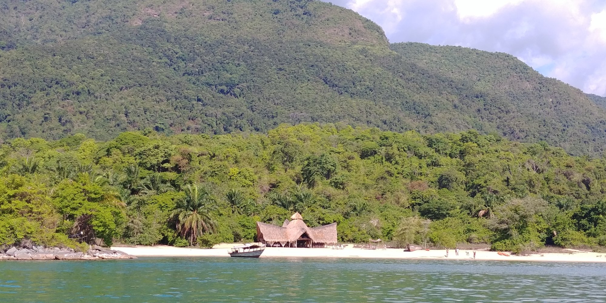 Forested Mahale mountain rising above a beach camp with guests on the white sand