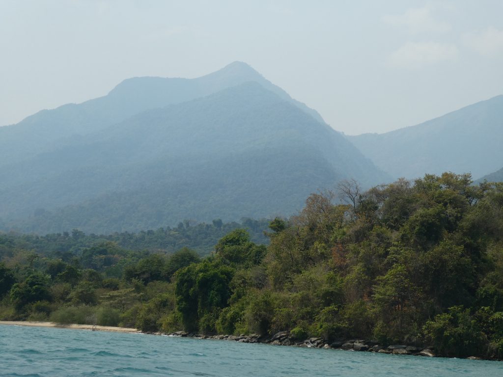 Hazy forested peak above rocky shoreline from turquoise water, Mahale Mountains National Park, Tanzania