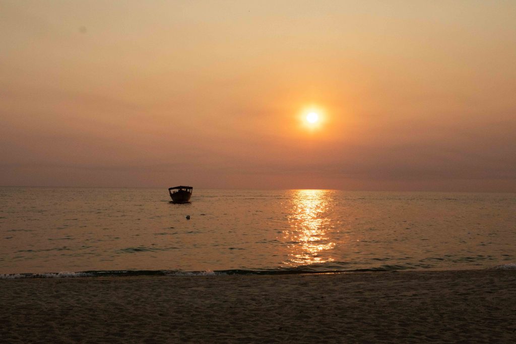 Hazy orange sun over lake with lone boat and empty sand, Mahale Mountains National Park, Tanzania