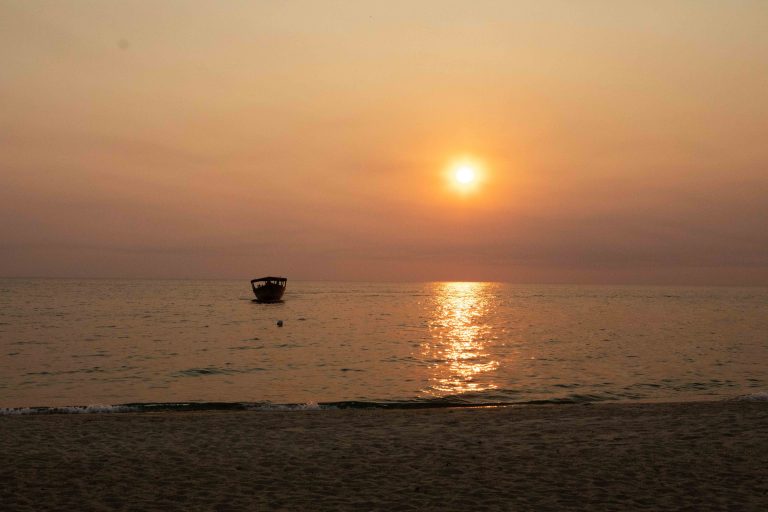 Hazy orange sun over lake with lone boat and empty sand, Mahale Mountains National Park, Tanzania